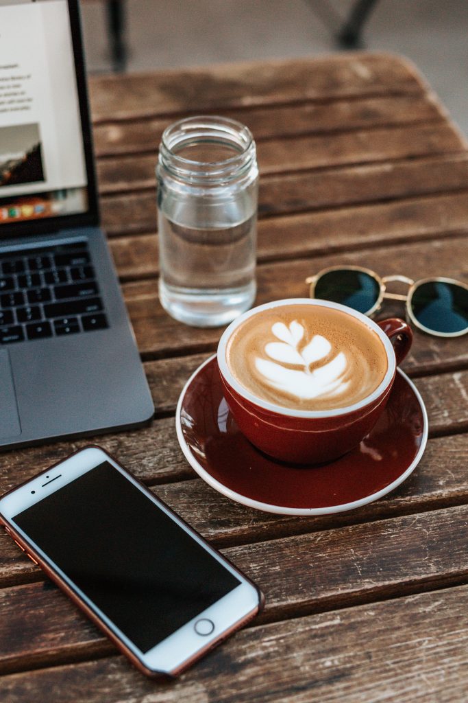 Coffee, sunglasses, water, laptop, and cellphone on wood table.